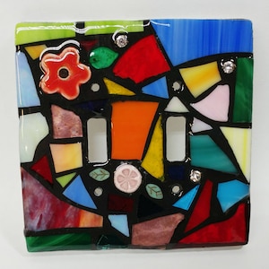 May include: A colorful mosaic light switch cover with a red, orange, yellow, green, blue, and pink design. The cover has a black border and two white switch plates.