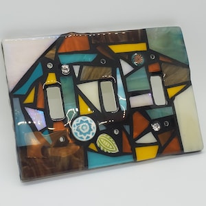 Könnte beinhalten: Ein bunter Mosaik-Lichtschalter mit geometrischem Design aus türkisfarbenen, braunen, gelben und orangefarbenen Glasstücken. Die Abdeckung hat zwei Schalteröffnungen und ist mit kleinen Strasssteinen verziert.