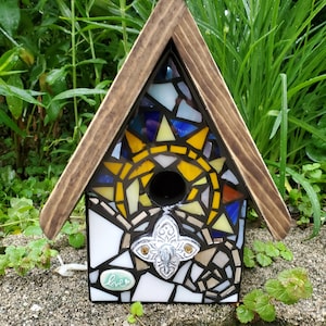 May include: A mosaic birdhouse with a wooden roof and a sun design. The birdhouse has a small hole for birds to enter and a small sign that says "love".