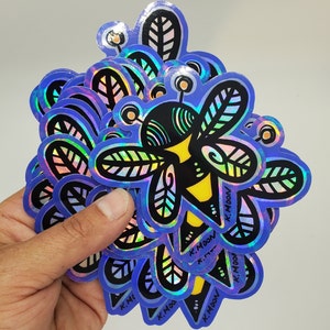 May include: A hand holds a stack of holographic bee stickers. The stickers have a blue iridescent sheen with a black and yellow bee design. The bee has leaf-shaped wings and the text "K. Moon" is printed on the stickers.