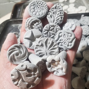 Pode incluir: Uma coleção de carimbos de argila cinza com vários desenhos, incluindo uma estrela, um redemoinho, uma folha e uma flor. Os carimbos são segurados em uma mão.