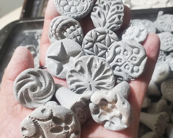 May include: A collection of grey clay stamps with various designs, including a star, a swirl, a leaf, and a flower. The stamps are held in a hand.