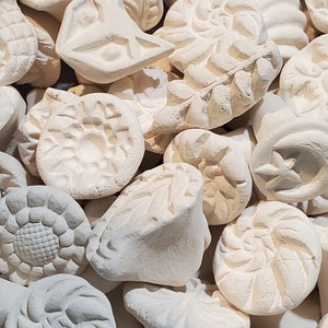 Pode incluir: Uma coleção de carimbos de cerâmica branca com vários desenhos, incluindo flores, folhas e padrões geométricos. Os carimbos são usados para criar impressões únicas em argila ou outros materiais.