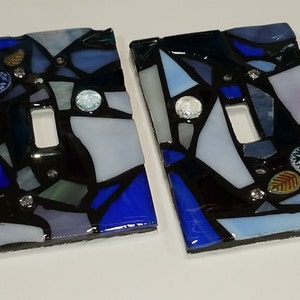 May include: Two blue and white mosaic light switch covers with black grout and decorative accents.