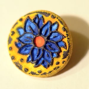 以下が含まれることがあります： 黄色い背景に青い花が描かれた丸いセラミックボタン。花の中心はオレンジ色です。