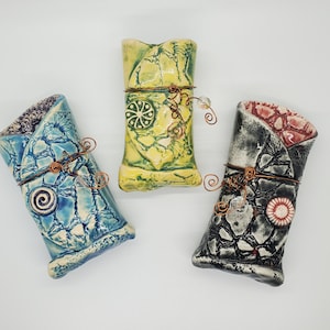 Op de afbeelding: Drie keramische vazen met een getextureerde, gebarsten afwerking. Elke vaas is versierd met een andere kleur glazuur en een uniek ontwerp. De vazen zijn gebonden met koperdraad en hebben heldere glazen kralen die eraan hangen.