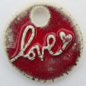 May include: A round ceramic charm with a white glaze and a red background. The word "love" is written in white with a heart at the end. The charm has a hole at the top for hanging.