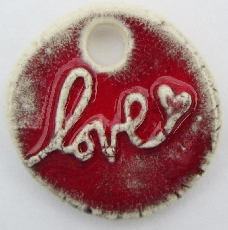 May include: A round ceramic charm with a white glaze and a red background. The word "love" is written in white with a heart at the end. The charm has a hole at the top for hanging.