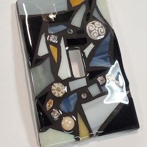 Könnte beinhalten: Ein schwarzer Lichtschalter-Abdeckung mit einem Mosaik-Design aus weißen, blauen, gelben und goldenen Glasstücken. Das Mosaik enthält kleine dekorative Elemente wie Strasssteine und ein Wirbeldesign.