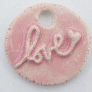 May include: A pink ceramic charm with a raised white inscription that reads "love" with a heart at the end. The charm has a hole for hanging.