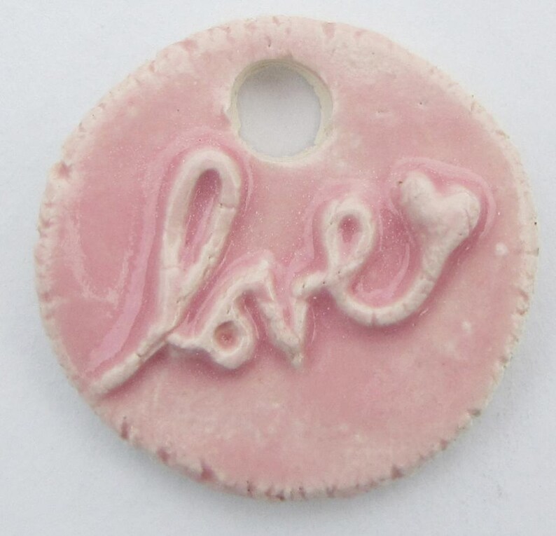 May include: A pink ceramic charm with a raised white inscription that reads "love" with a heart at the end. The charm has a hole for hanging.