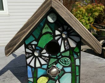May include: A mosaic birdhouse with a brown wooden roof. The birdhouse is decorated with green and blue glass tiles in a floral pattern. The birdhouse has a small hole for birds to enter.
