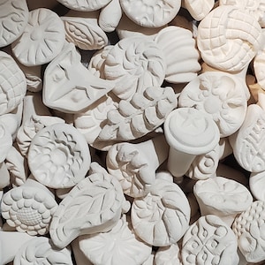 May include: A collection of white ceramic stamps with various designs, including floral, geometric, and abstract patterns. The stamps are arranged in a random order, creating a visually interesting texture.