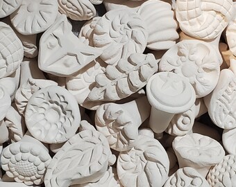 May include: A collection of white ceramic stamps featuring a variety of designs, including floral, geometric, and abstract patterns. The stamps are arranged randomly, creating a visually interesting texture.