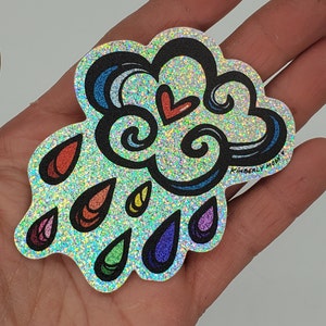 May include: A sparkly silver sticker featuring a blue and white cloud with a red heart and rainbow raindrops. The sticker reads "Kimberly Moon" at the bottom.