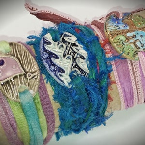 May include: Three colourful bracelets with ceramic charms. The charms are decorated with blue, green, and purple paint and have a textured surface. The bracelets are made of fabric and have a frayed edge.