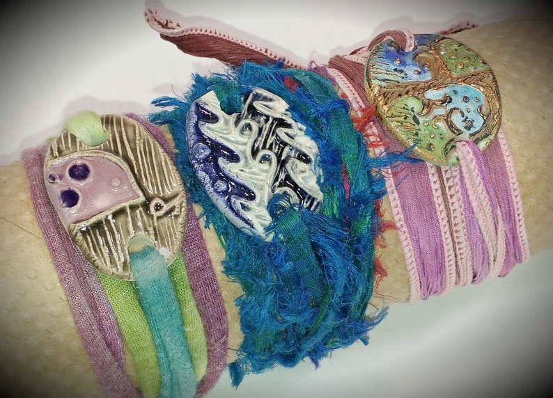 May include: Three colourful bracelets with ceramic charms. The charms are decorated with blue, green, and purple paint and have a textured surface. The bracelets are made of fabric and have a frayed edge.