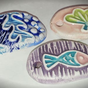 May include: Three ceramic ornaments with a sea life theme. One ornament is blue and white with a wave design, another is pink with a green seashell, and the last is purple with a turquoise fish.