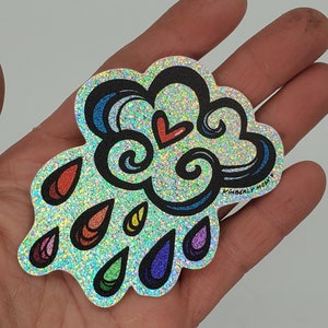 May include: A holographic glitter sticker featuring a rainbow cloud with a red heart and raindrops. The sticker is designed by Kimberly Moon.