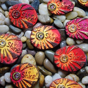 May include: A collection of oval-shaped, hand-painted ceramic ornaments with a sunburst design. Each ornament features a gold-colored eye with a rhinestone center and the word "Shine!" in gold lettering.