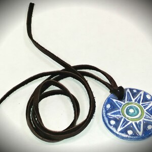 May include: A blue and white ceramic pendant with a starburst design and a brown leather cord. The pendant has a green and blue eye in the centre.