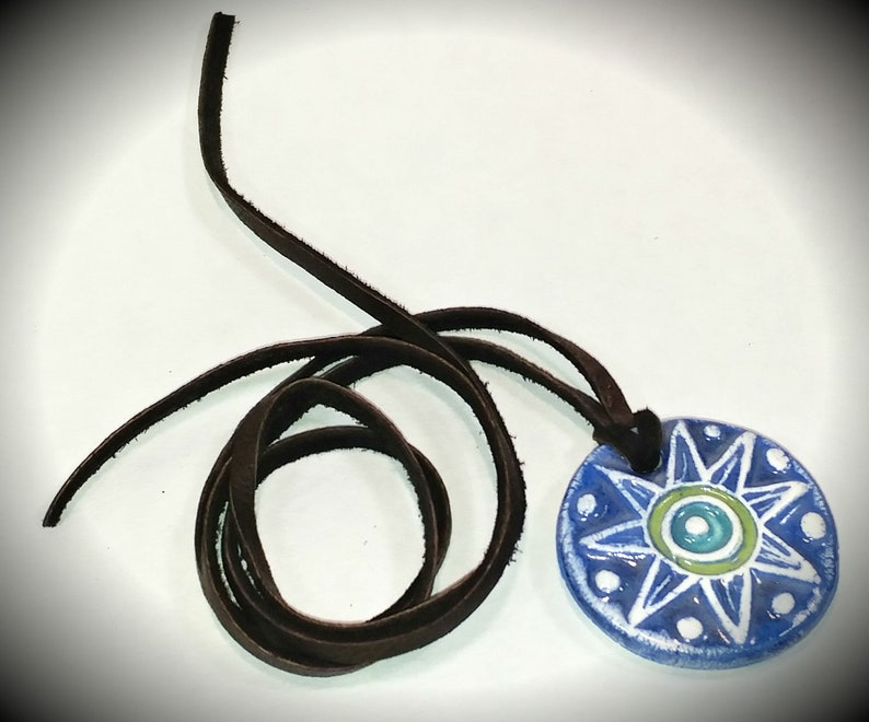 May include: A blue and white ceramic pendant with a starburst design and a brown leather cord. The pendant has a green and blue eye in the centre.