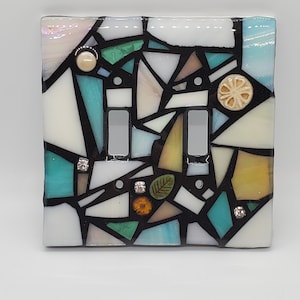 May include: A mosaic light switch cover with a geometric design in shades of white, blue, green, and yellow. The cover features small decorative accents, including a flower, a leaf, and rhinestones.