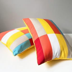 May include: Two colorful throw pillows with a patchwork design. The pillows feature a vibrant mix of colors, including red, orange, yellow, white, and blue. The pillows are rectangular and have a yellow border.