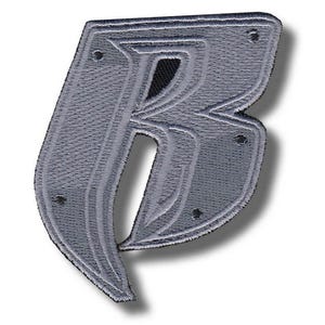 以下が含まれることがあります： 文字「B」の形をしたグレーの刺繍パッチ。パッチは、一連の平行線と小さな暗いリベットを備えたテクスチャの外観をしています。文字はより濃いグレーで縁取られています。