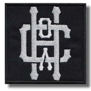 May include: A square, black embroidered patch with a white monogram design. The monogram features the letters "HAC" in an ornate, stylized font. The patch has a black border and a textured appearance.