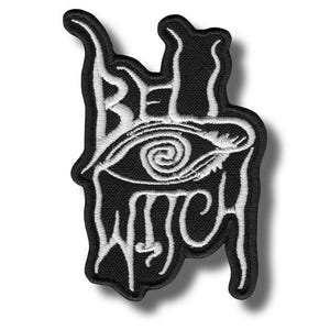 Può includere: Toppa nera con testo ricamato bianco e disegno di un occhio. La toppa recita "BE WITCH" in un font stilizzato, con l'occhio al centro. La toppa ha un bordo nero ed è probabilmente una toppa da stirare o da cucire.