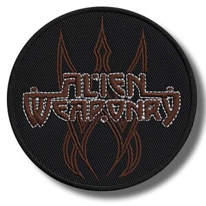 May include: A round, black patch with the words "ALIEN WEAPONRY" in a brown, stylized font. The text is outlined in white stitching. Brown, flame-like designs are above and below the text, also outlined in white. The patch is likely for a jacket or bag.
