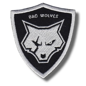 May include: Embroidered patch featuring a shield design with a black background and a white wolf head. The text "BAD WOLVES" is arched above the wolf. The patch has a white border and is likely for clothing or accessories.
