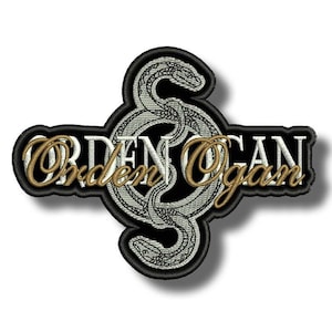 Peut inclure: Écusson brodé avec les mots "Orden Ogan" en lettres dorées sur un motif de serpent argenté. Le patch a une bordure noire et un effet d'ombre, créant un aspect 3D. Le serpent est finement détaillé.