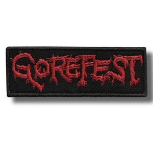 Puede incluir: Un parche rectangular negro con la palabra "GOREFEST" bordada en rojo. La tipografía es estilizada. El parche tiene una textura, con el hilo rojo creando un efecto en relieve sobre el fondo negro. Para ropa o accesorios.