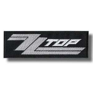 May include: A rectangular black patch with the words "ZZ TOP" embroidered in white. The patch has a simple, clean design with a shadow effect, suitable for jackets, bags, or other apparel.