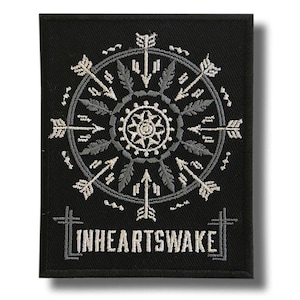 May include: Black embroidered patch with a circular design of arrows, feathers, and a central floral pattern. The text "INHEARTSWAKE" is embroidered at the bottom. The patch is rectangular in shape.