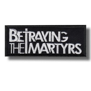 Può includere: Una toppa rettangolare nera con testo ricamato bianco che recita "BETRAYING THE MARTYRS". La toppa ha un bordo nero e un effetto ombra, suggerendo che sia in rilievo.