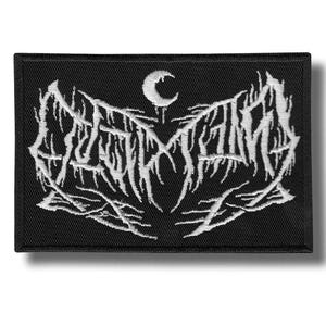 Pode incluir: Patch retangular preto com texto bordado branco e uma lua crescente. O texto, em uma fonte gótica, é provavelmente o nome de uma banda. O patch tem uma borda preta e provavelmente é feito de tecido.