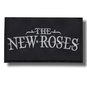 Puede incluir: Parche rectangular negro con el texto bordado "THE NEW ROSES" en blanco. El texto está en una fuente estilizada. El parche tiene un borde negro y es para ropa o accesorios.