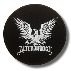 Peut inclure: Un écusson rond noir avec un motif brodé blanc d'un oiseau aux ailes déployées. Le texte "ALTER BRIDGE" est brodé en blanc en dessous de l'oiseau.