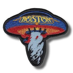 Op de afbeelding: Geborduurde patch met het woord "BOSTON" in gouden letters, tegen een blauwe en rode achtergrond. De patch heeft een unieke vorm met een lichtblauw wezenontwerp aan de onderkant, omlijnd in zwart.