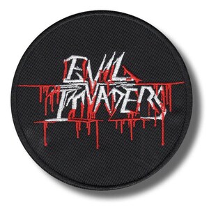 Può includere: Una toppa rotonda e nera ricamata con le parole "EVIL INVADERS" in bianco, con dettagli rossi gocciolanti. La toppa è probabilmente per una giacca o una borsa e ha un tema heavy metal.