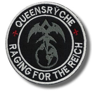 May include: A round, black patch with white embroidered text and a gray emblem. The text reads "QUEENSRYCHE" at the top and "RAGING FOR THE REICH" along the bottom. Two red crosses flank the emblem.