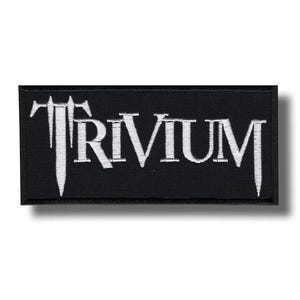 Puede incluir: Parche rectangular negro con la palabra "TRIVIUM" bordada en blanco. Las letras tienen una fuente gótica estilizada. El parche es probablemente para una banda o merchandising musical.