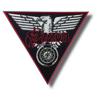 Op de afbeelding: Een zwarte, driehoekige patch met een rode rand. De patch heeft een witte adelaar met uitgespreide vleugels boven het rode woord "SAXON". Daaronder bevindt zich een wit en zilver cirkelvormig ontwerp. Deze patch is waarschijnlijk voor een jas of tas.