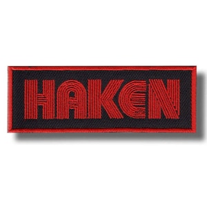 May include: Embroidered patch with the word "HAKEN" in red, stylized lettering. The text is on a black background with a red border. The patch is rectangular and likely designed to be sewn or ironed onto fabric.