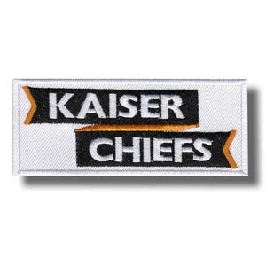 Puede incluir: Parche bordado con las palabras "KAISER CHIEFS" en letras blancas sobre fondo negro. El parche tiene un borde blanco y detalles en naranja. El parche es rectangular.