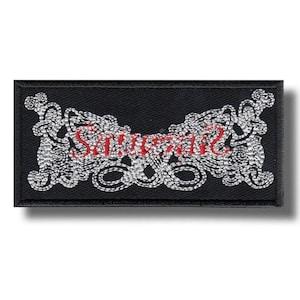 Puede incluir: Parche rectangular negro con la palabra "Savage" bordada en rojo, rodeada de bordados plateados ornamentados. El parche es probablemente para ropa o accesorios.