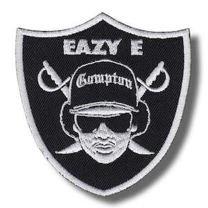 Può includere: Toppa ricamata in bianco e nero a forma di scudo. La toppa presenta il testo "EAZY E" in alto, con un'immagine stilizzata di una persona con occhiali da sole e un cappello Compton. Due spade incrociate sono su entrambi i lati dell'immagine.